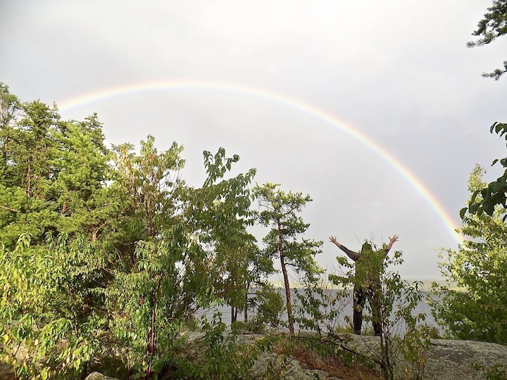 woman standing with arms upraised under a rainbow over a lake