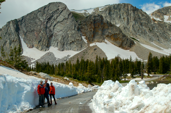 four people standing on a road with high snowbanks, mountains directly behind