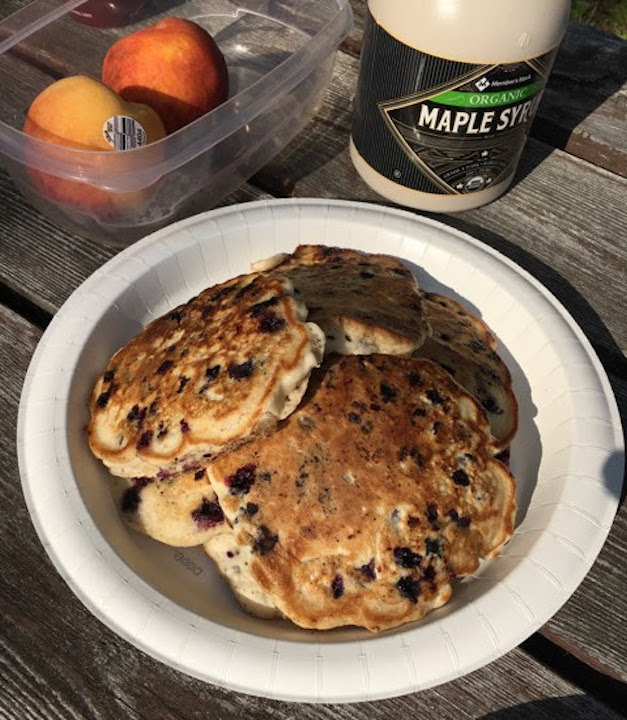 a plate of blueberry pancakes, along with maple syrup and fresh peaches