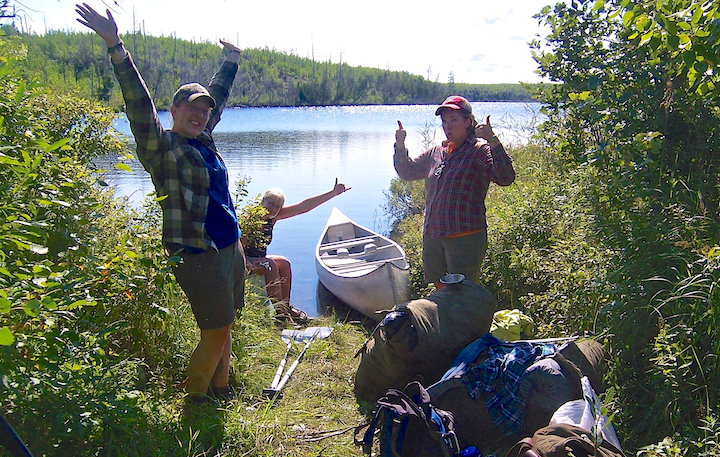 Three women with thumbs up, with their canoe and gear at a portage