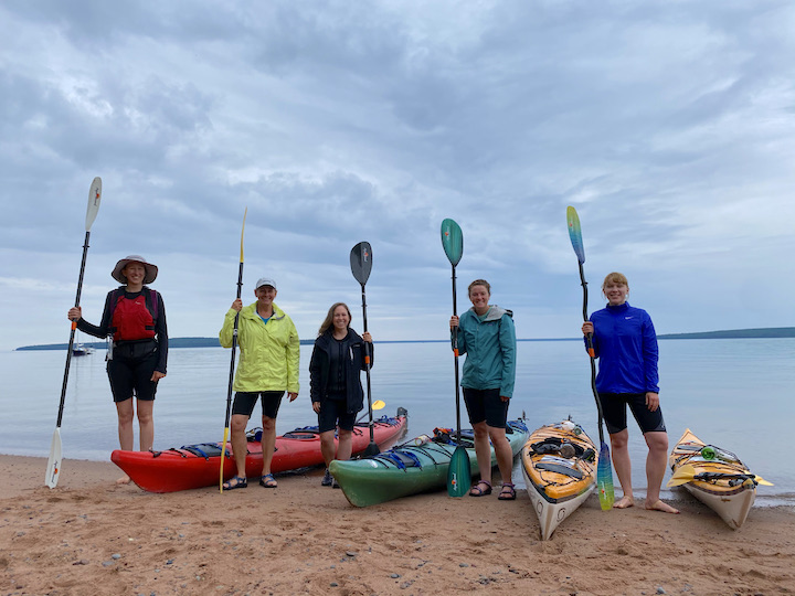 five women of various ages on a sand beach with their kayaks and paddles; Lake Superior behind them and the mainland on the horizon