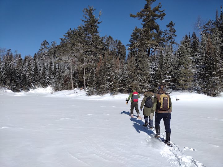 three snowshoers hiking across a frozen lake towards some trees