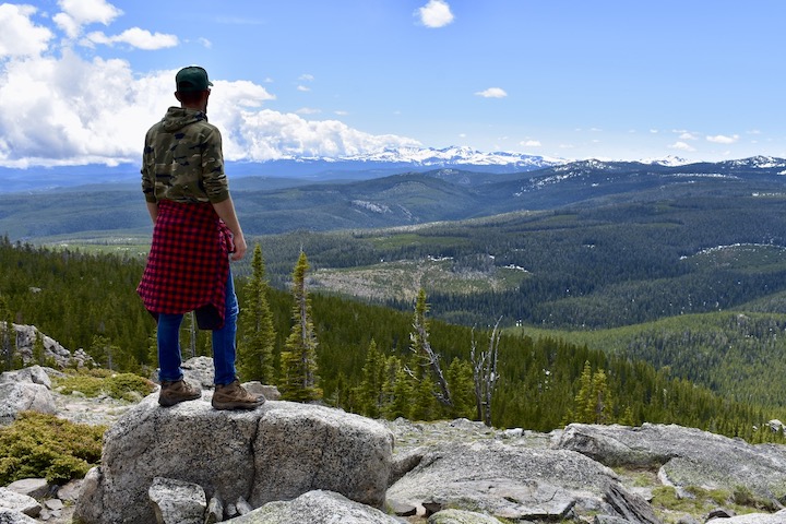 young man stands on a boulder overlooking miles of forest and mountains
