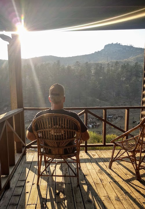 man sits on a cabin porch overlooking a mountain valley in early evening