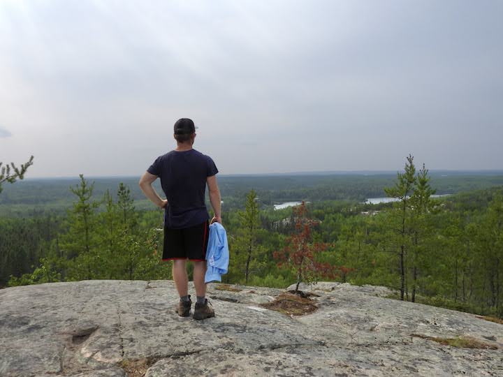 man stands on a granite high point overlooking forests and lakes