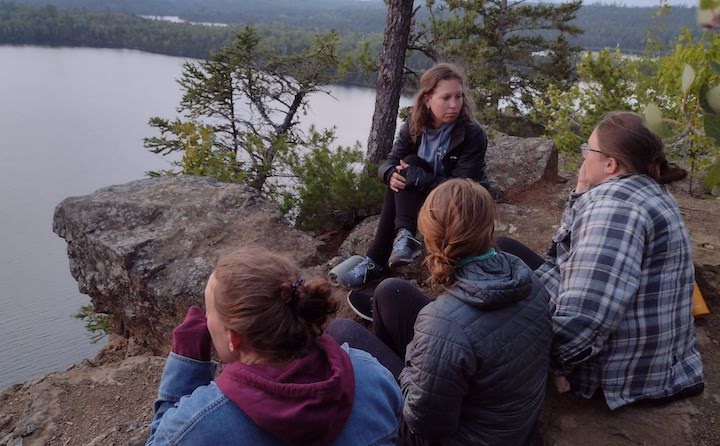 four women talk together on a cliff high up over a lake