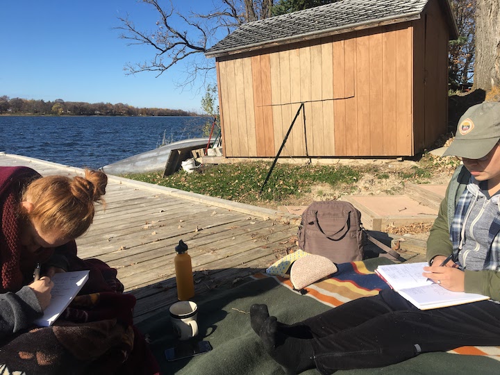 two women sit on a lakeside dock writing in journals