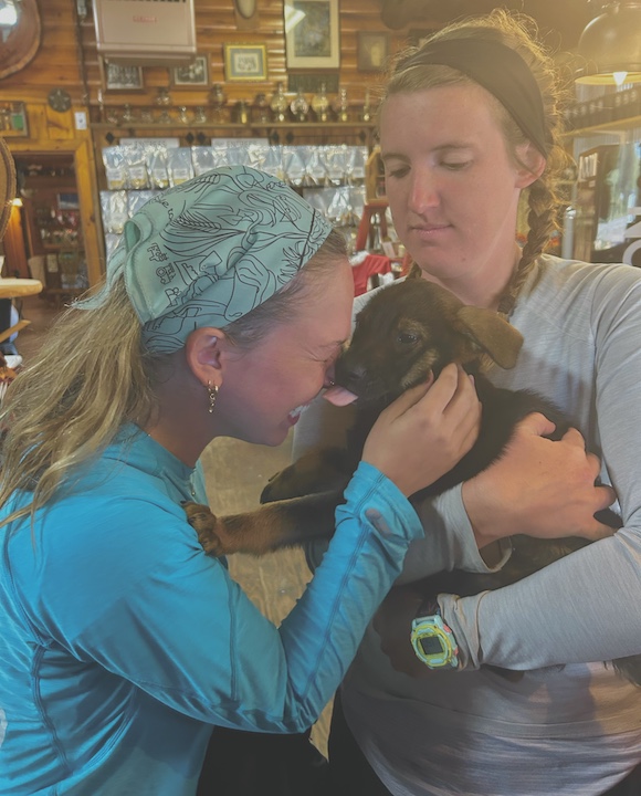 one woman holds a puppy while another gets a puppy kiss