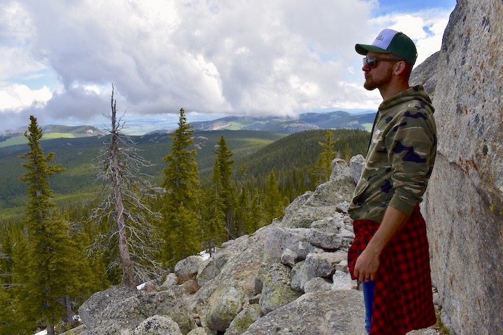 young man stands on a mountaintop overlooking more mountains and trees