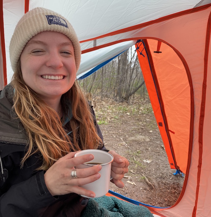 young woman drinks her morning coffee inside her tent in the woods
