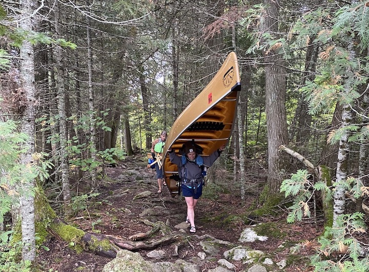 woman portages a canoe on a wilderness trail, followed by another portaging gear