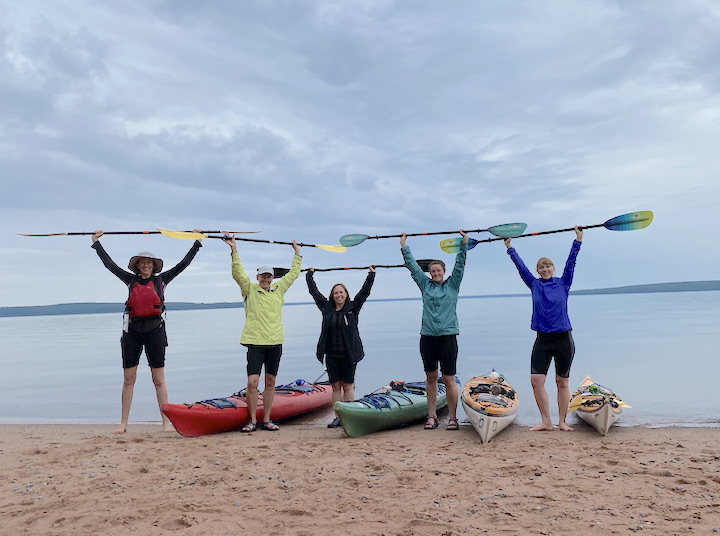 five women hold up kayak paddles while standing on shore next to their kayaks