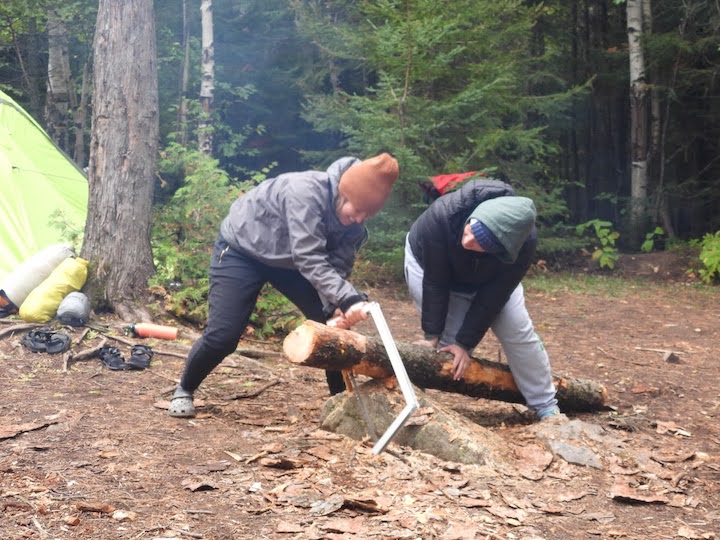 one woman saws through a log at a campsite while another holds the log
