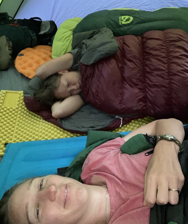 young woman sound asleep in her sleeping bag inside the tent; selfie taken by second woman, also in sleeping bag