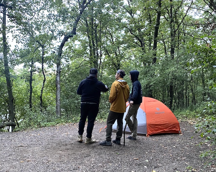 three men stand besides a tent in the campsite