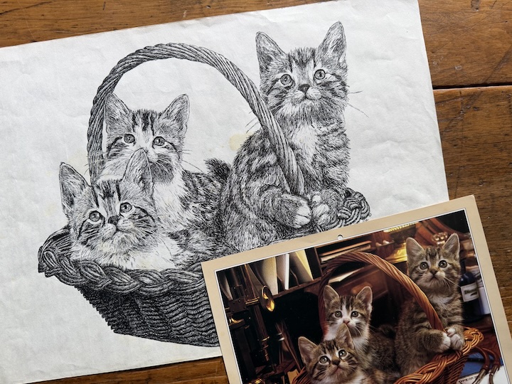pen and ink drawing of three kittens in a basket, with the source photograph laying next to it